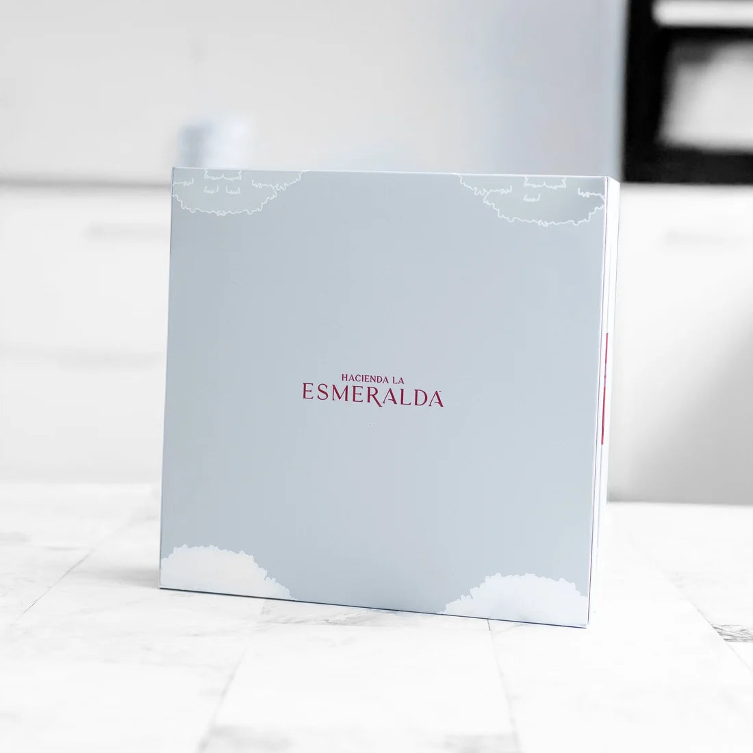 Hacienda La Esmeralda Geisha gift box closed, pale blue lid with dark red Esmeralda logo and cloud motif on marble