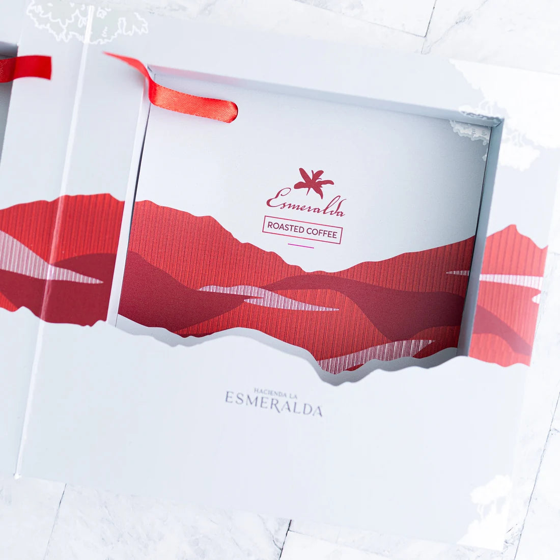 White coffee gift box with a red ribbon handle and a cutout window showing red mountain-style artwork. The front reads HACIENDA LA ESMERALDA.
